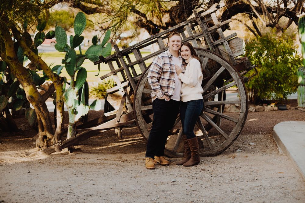 Two people pose together against a rustic wooden wheel in an outdoor garden setting.