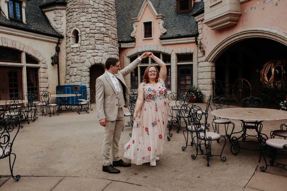 Couple dancing in front of a castle-like venue, man in tan suit spinning woman in floral wedding dress.