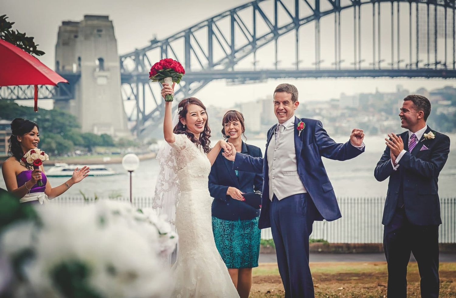 Joyful bride raises bouquet with groom and guests near Sydney Harbour Bridge after wedding ceremony.