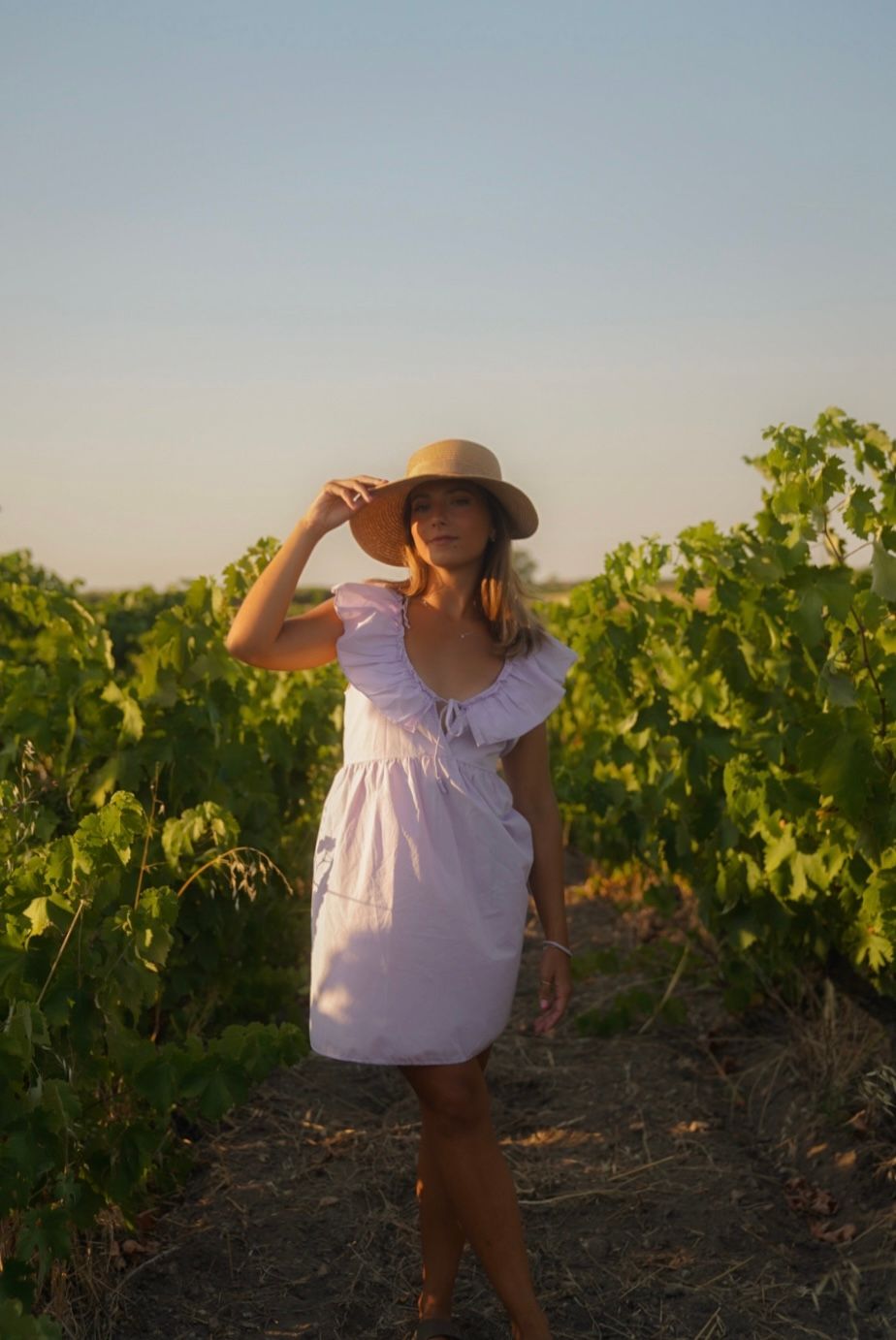Playful pose in white dress and sun hat between vineyard rows.