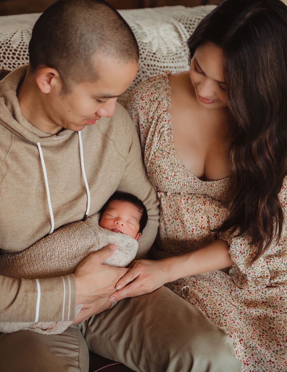 Mom and dad holding their newborn baby sitting on a couch in their house.