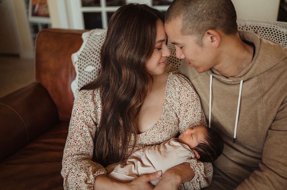 Mom and dad holding their newborn baby sitting on a couch in their house.