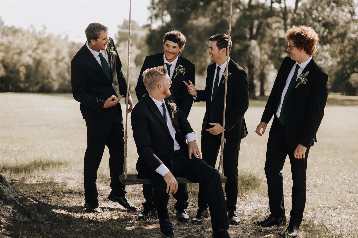 A group of groomsmen in black suits share a candid moment outdoors during a wedding photoshoot.