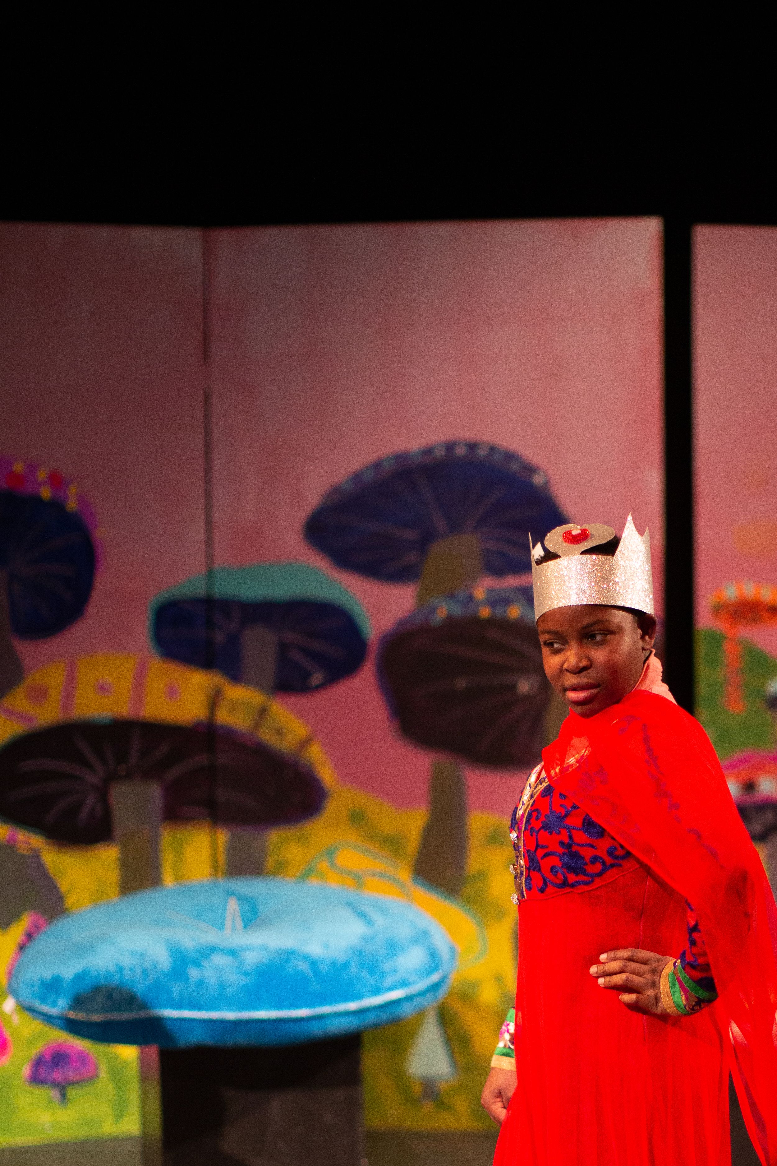 Performer in red cape and crown poses against colorful mushroom-themed stage backdrop.