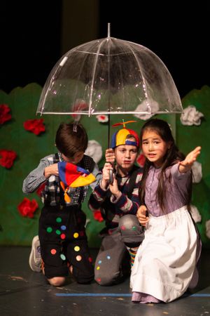Young performers crouch under clear umbrellas during a stage production with colorful props.