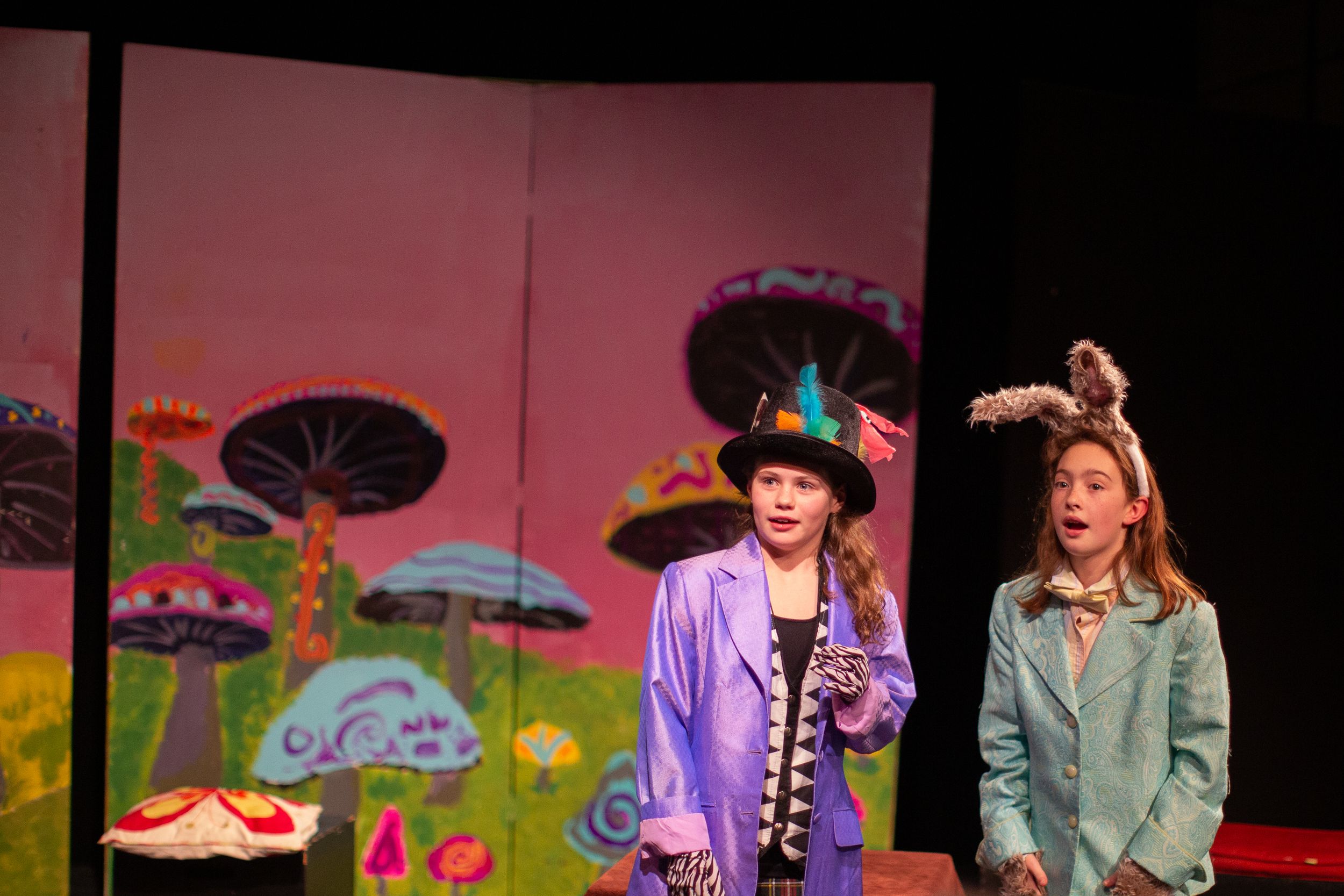 Stage performers in costume act against a whimsical backdrop with painted mushrooms.