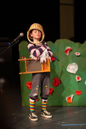Small performer in striped socks and winter hat stands on stage with green backdrop and red decorations.