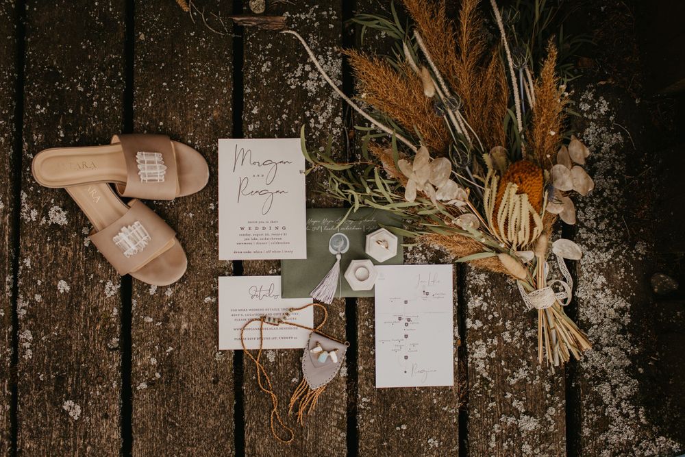 Wedding details flatlay with invitation, dried flowers and nude shoes on rustic wooden surface.