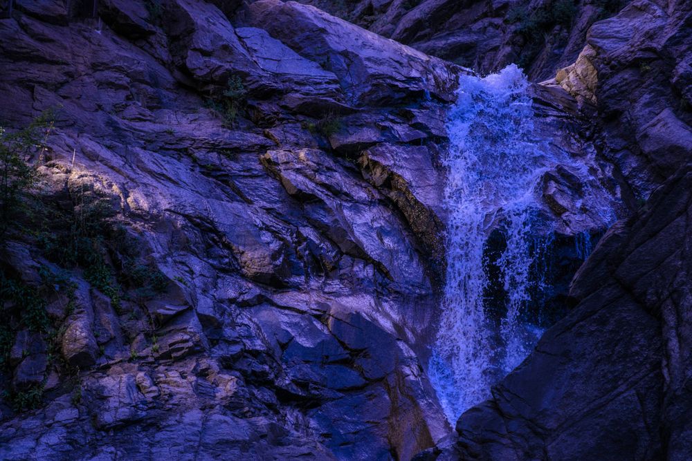 Dramatic blue waterfall illuminated against dark rocky cliffs in nighttime or low light conditions.