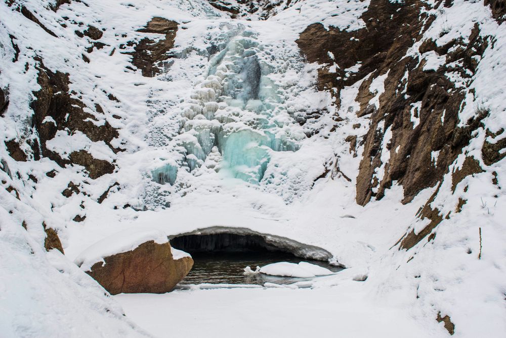 An icy blue frozen waterfall forms a natural arch in a snow-covered rocky landscape.