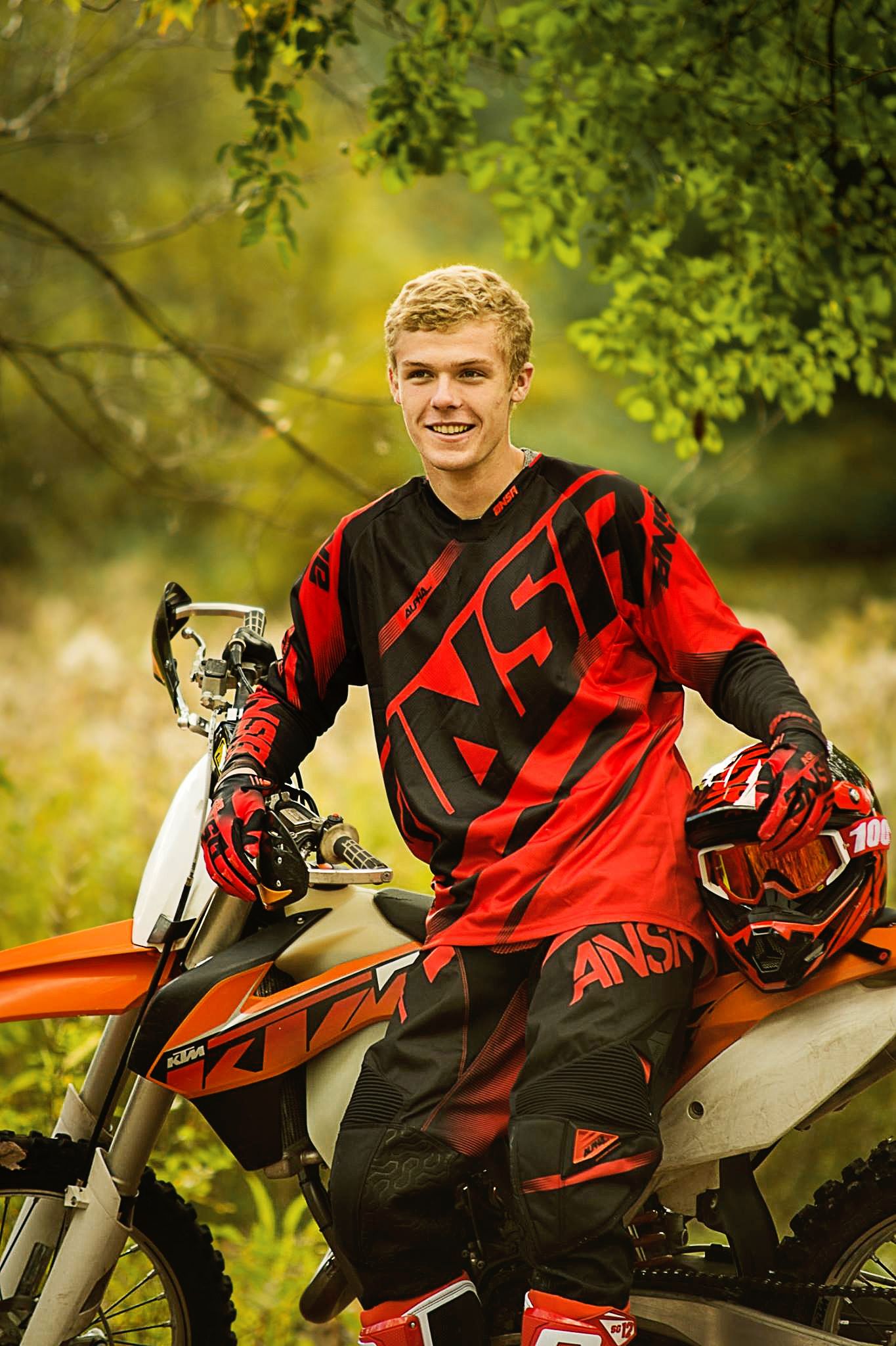 Dirt bike rider in red and black gear poses confidently on motorcycle against green foliage background.