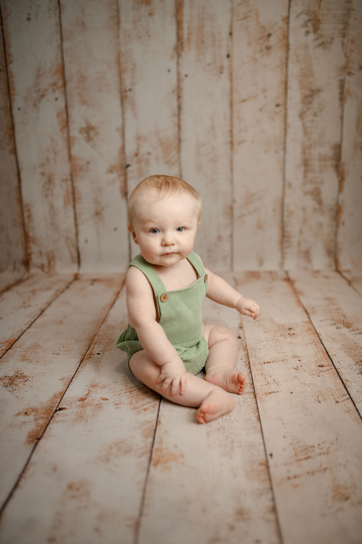 Baby in sage green romper sits on wooden floor against light backdrop.