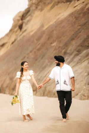A couple walks hand in hand along a sandy beach with cliffs in the background.