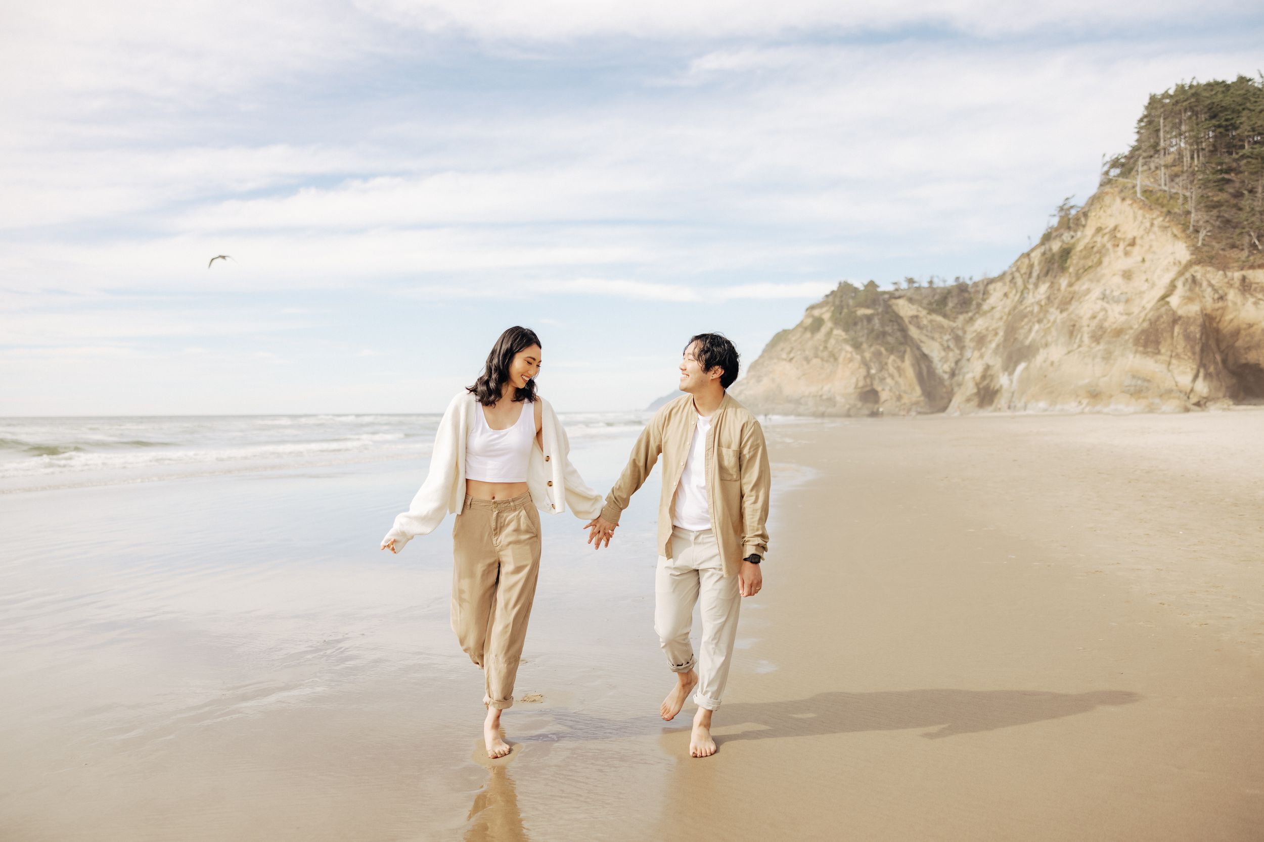 A couple walks hand in hand along a scenic beach with cliffs in the background.