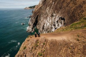 Two people walk along a dramatic coastal cliff trail overlooking the ocean.