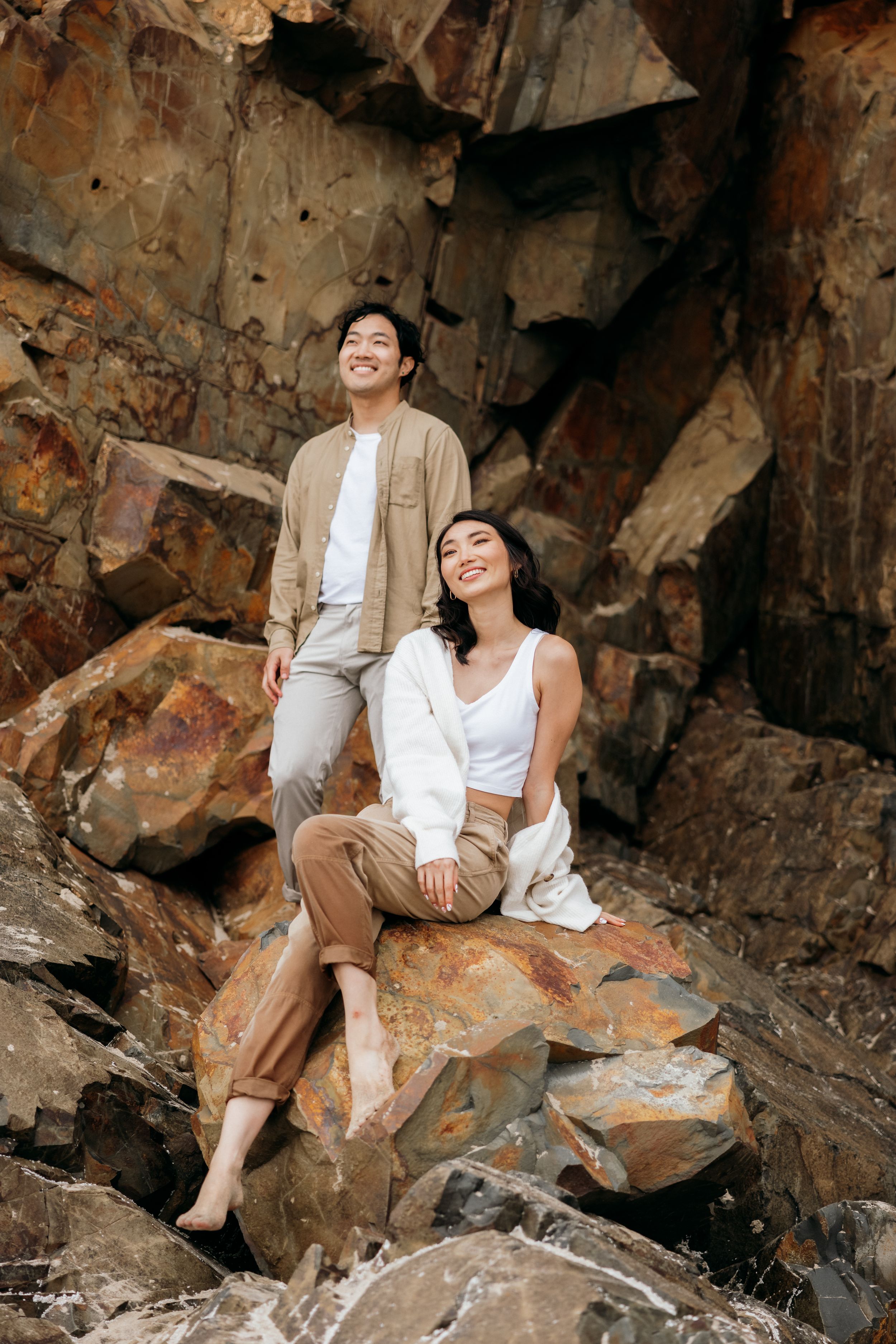 A couple poses casually on rocky coastal formations wearing neutral-toned outfits.
