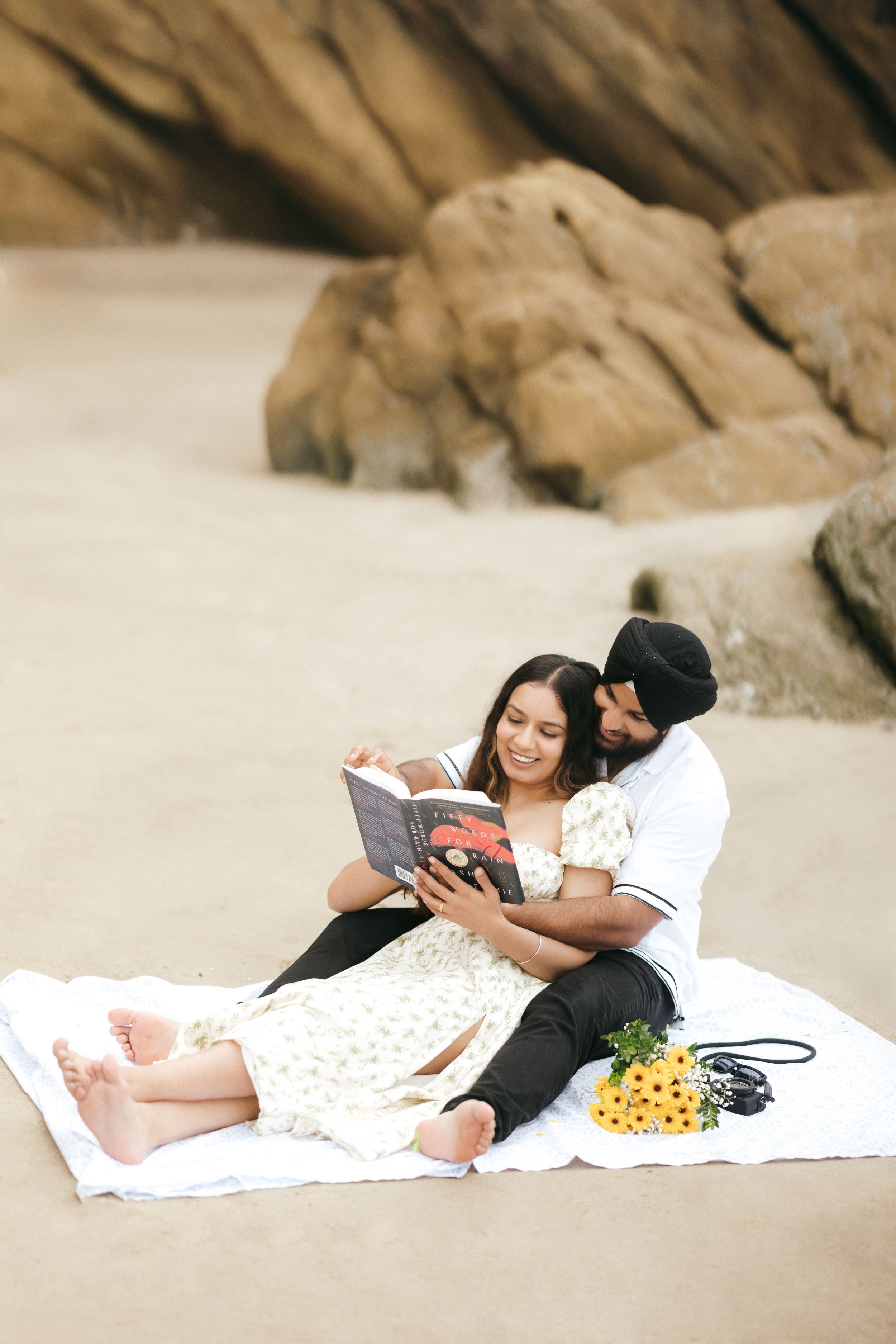 A couple reads together while having a picnic on a sandy beach near coastal rock formations.