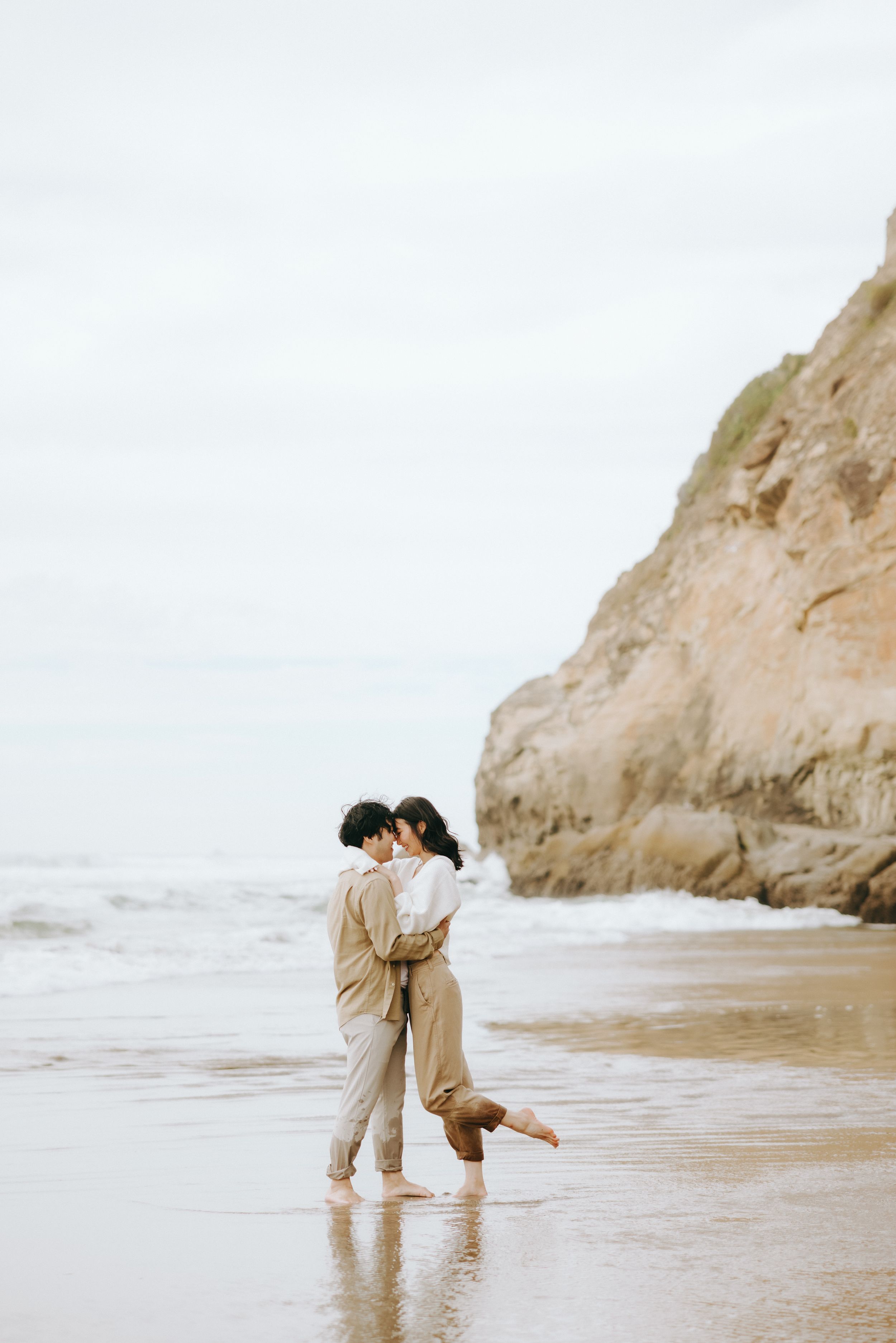 A romantic couple embraces on a misty beach near towering cliffs during a serene moment.