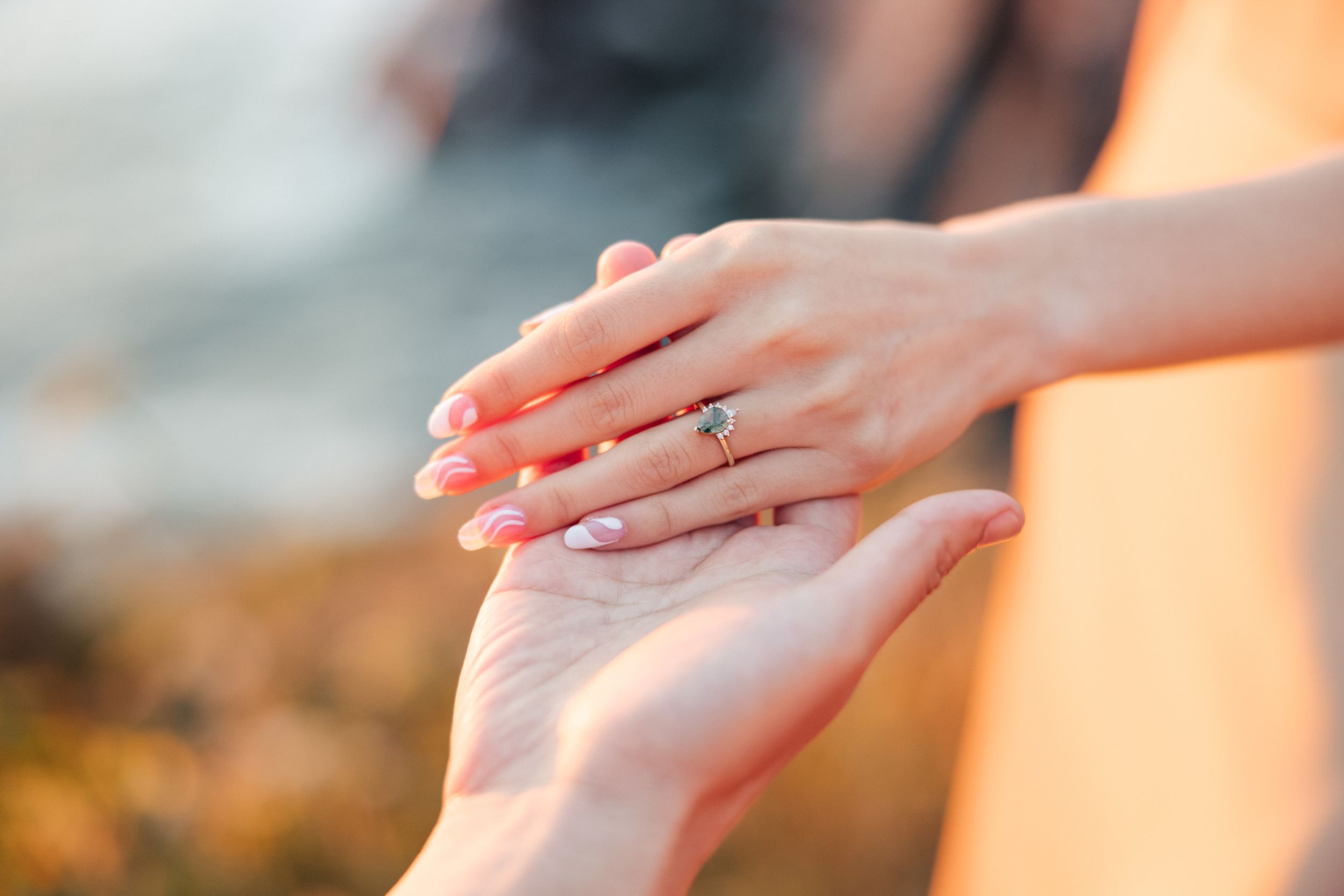 Close-up of hands with engagement ring touching in warm sunset light.
