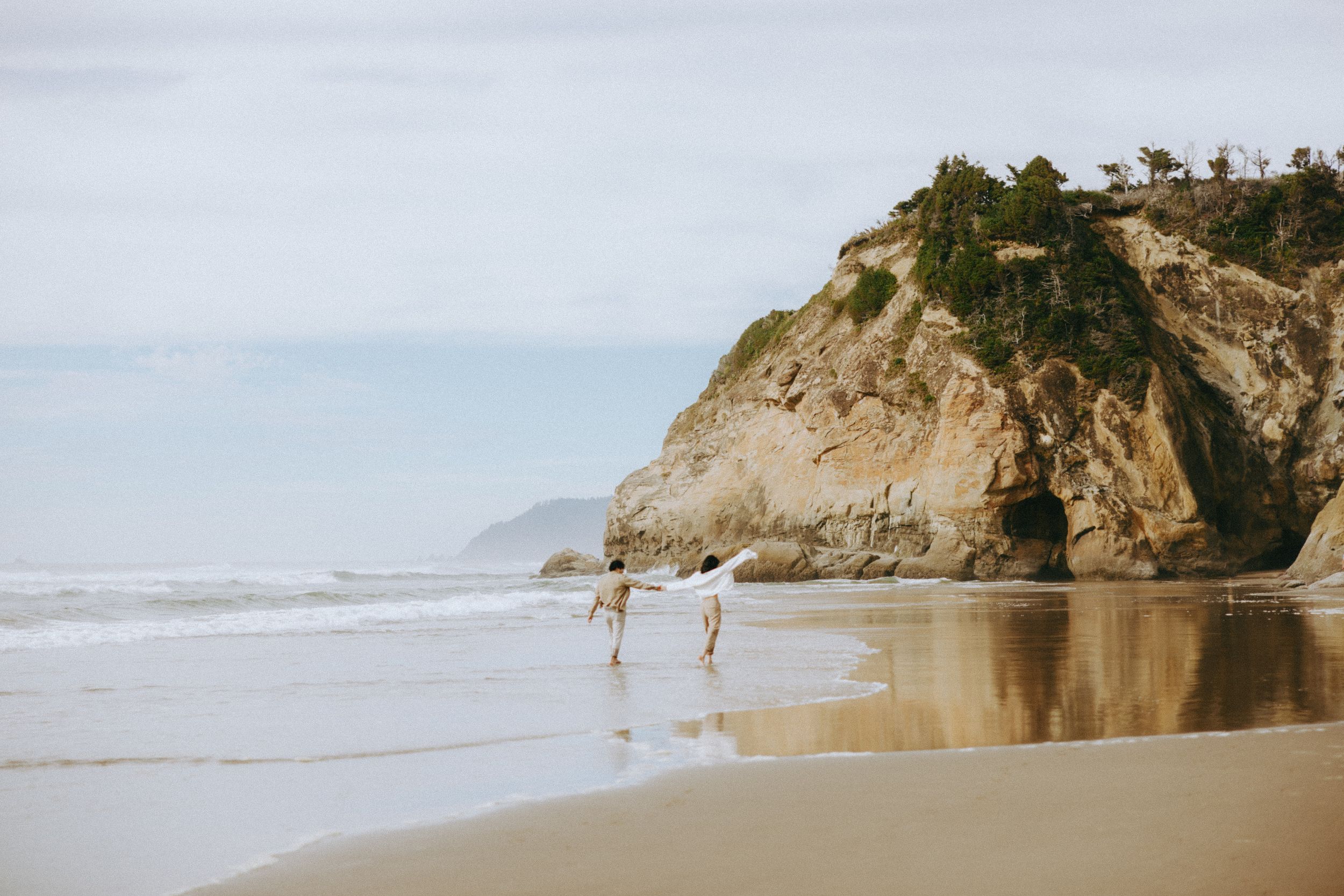 Two people walk along a dramatic coastline with towering cliffs and crashing waves.