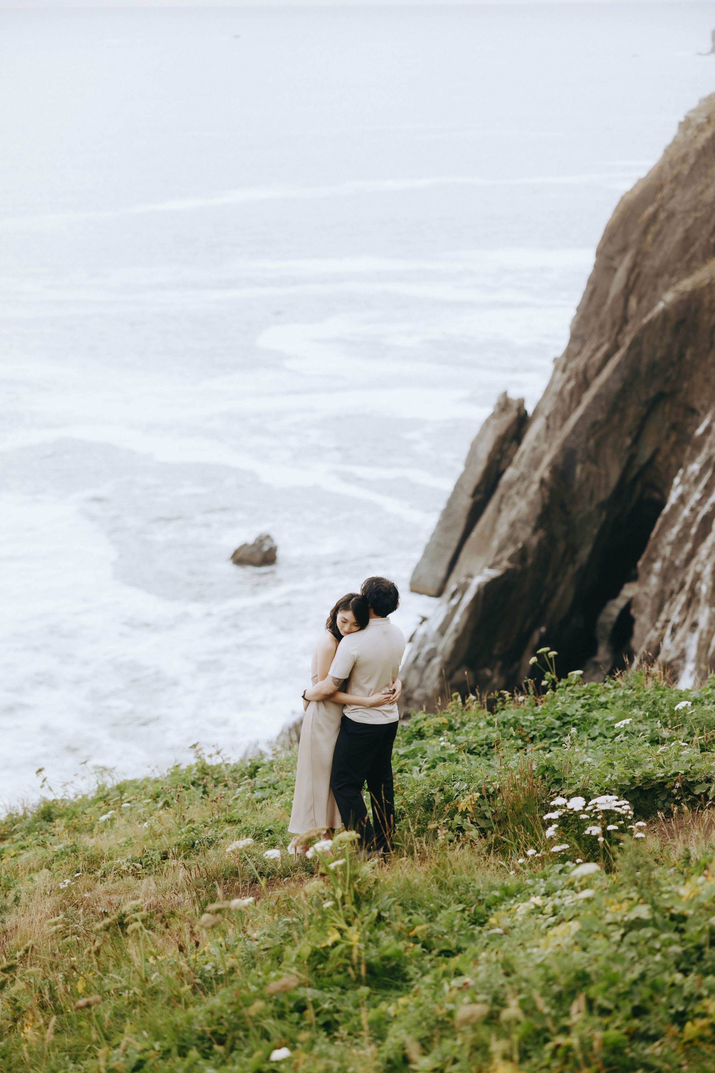 A couple shares an embrace on a grassy cliff overlooking the misty ocean.