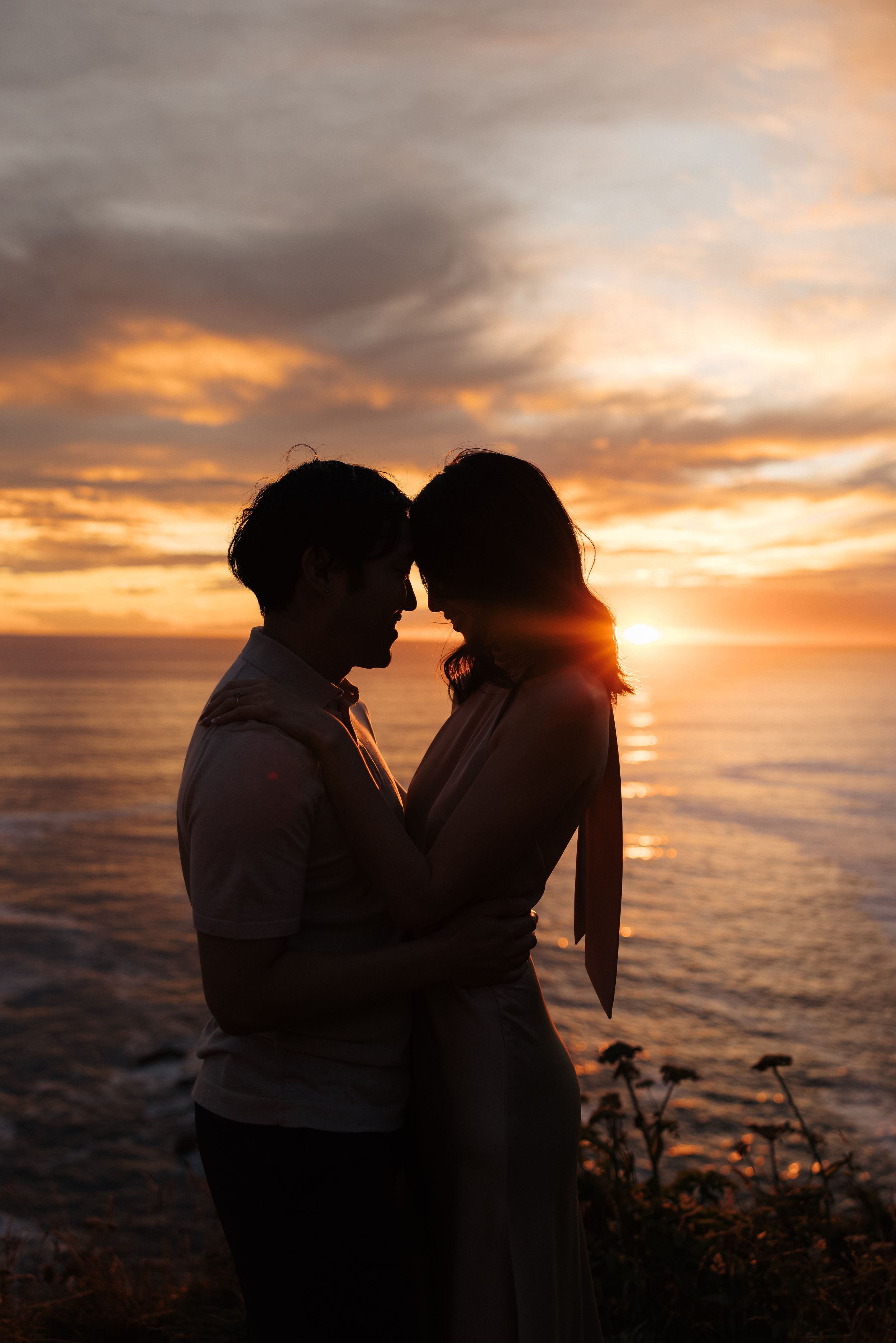 A silhouetted couple stands close together against a dramatic orange sunset over the ocean.