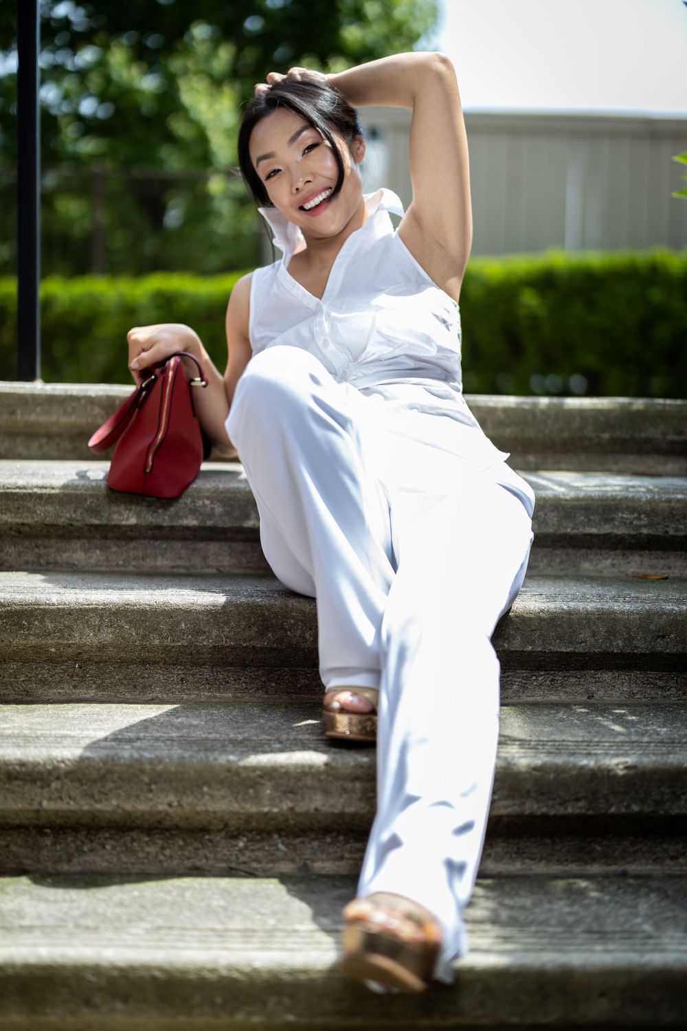 Casual portrait on concrete steps featuring white outfit and red handbag in outdoor setting.