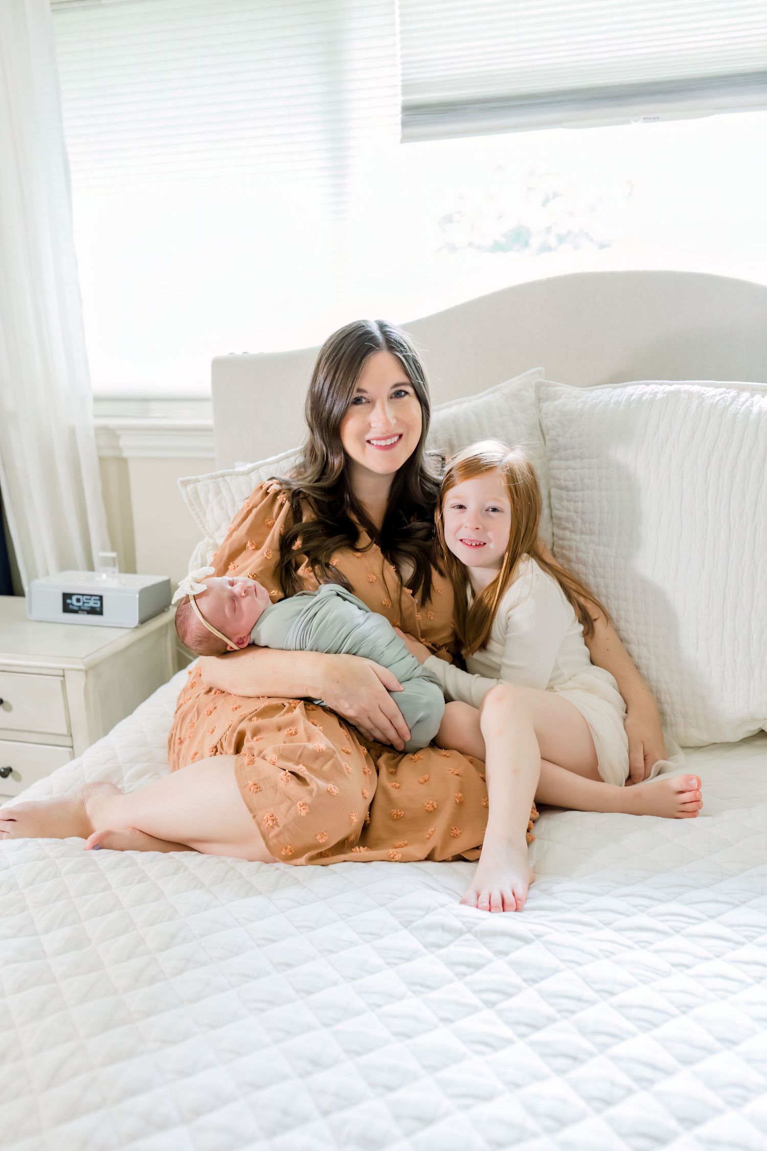 A woman in a peach dress sits on a white bed holding a baby with a young child beside her.