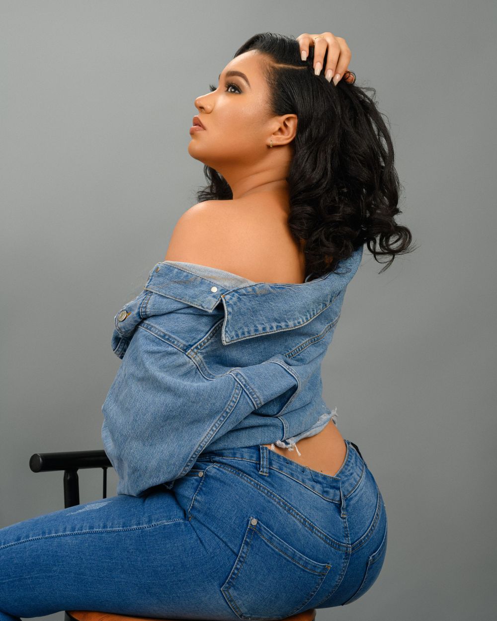 Fashion portrait of a person in denim wearing an off-shoulder jean jacket against a grey background.