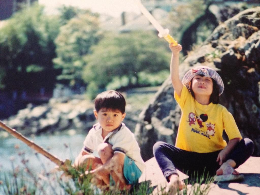 Two children fishing by a pond, one wearing a yellow Mickey Mouse t-shirt.