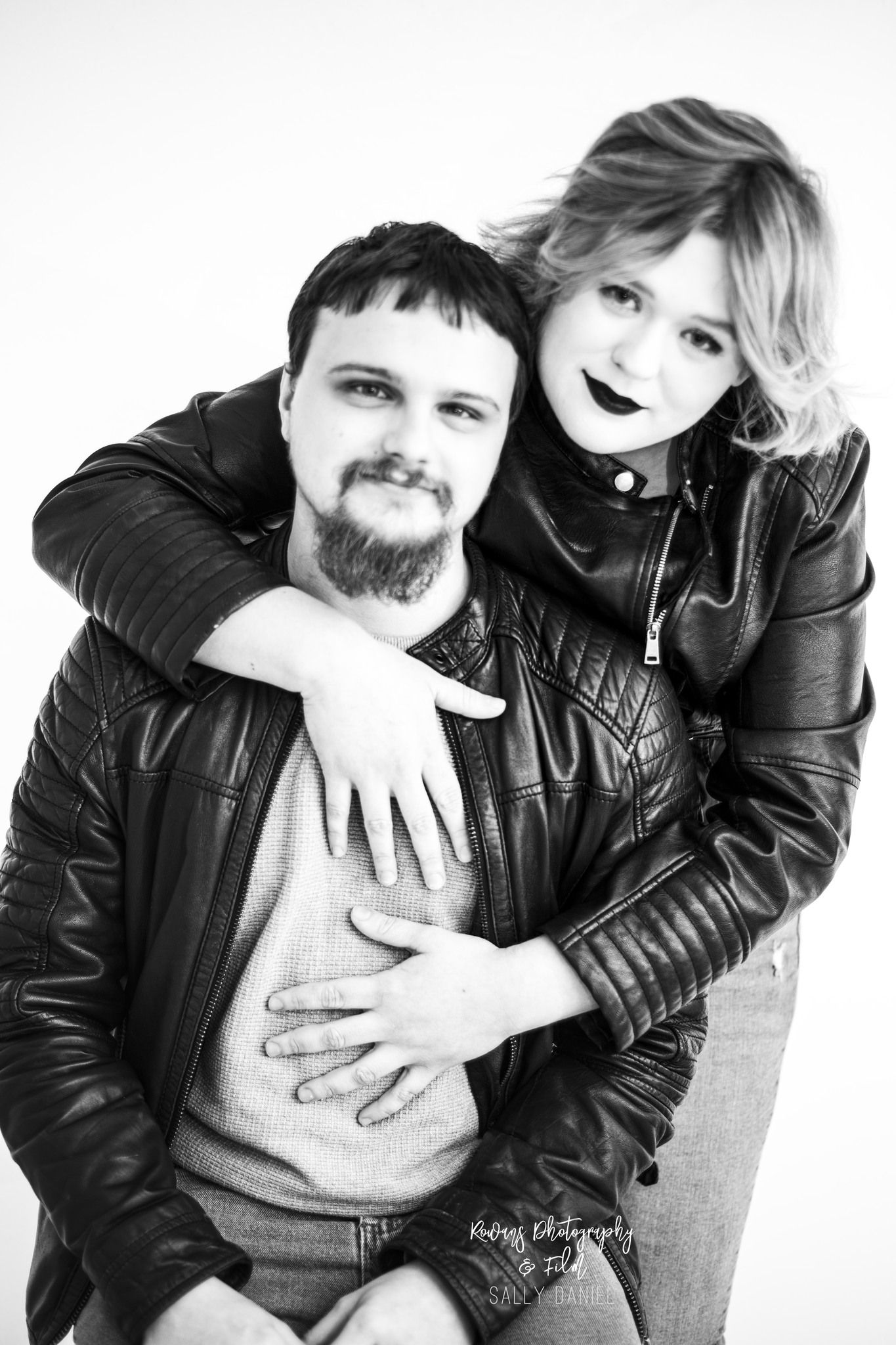 A black and white studio portrait of a couple in leather jackets embracing against white backdrop.