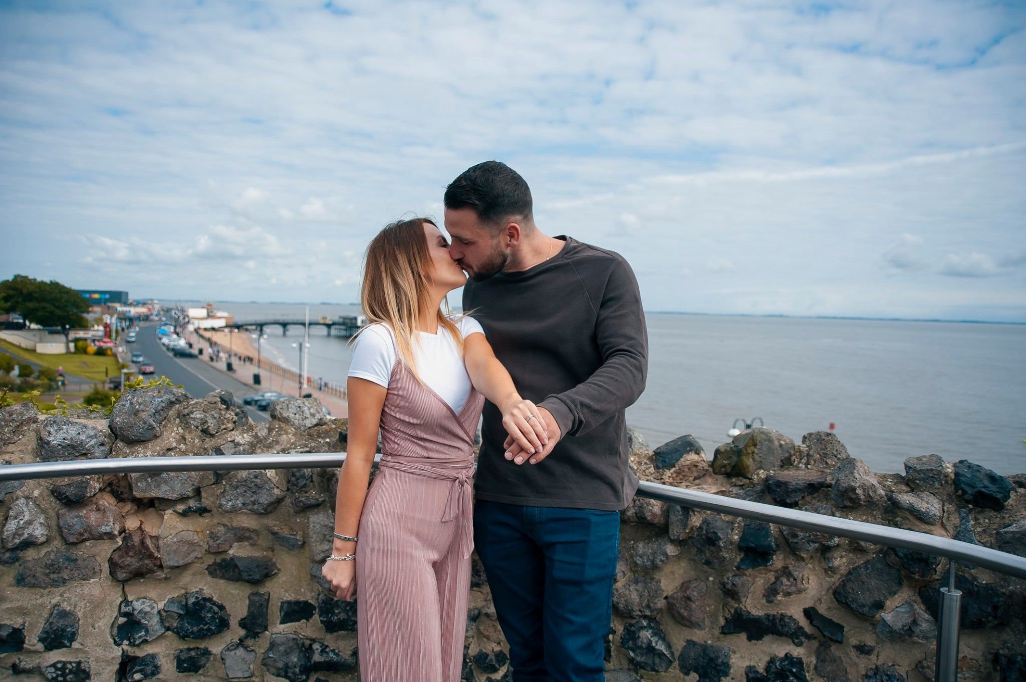 A couple sharing a tender moment overlooking a coastal view from a stone wall viewpoint.