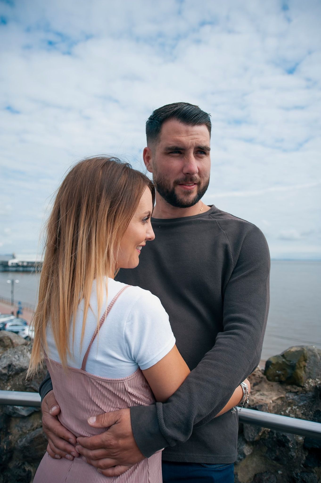A couple embracing on a coastal viewpoint with blue skies and ocean in the background.