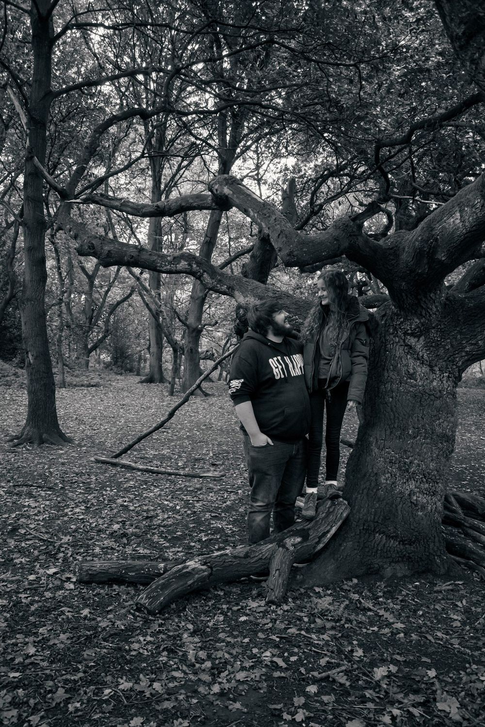 A couple posing together near a large tree in a moody black and white forest setting.
