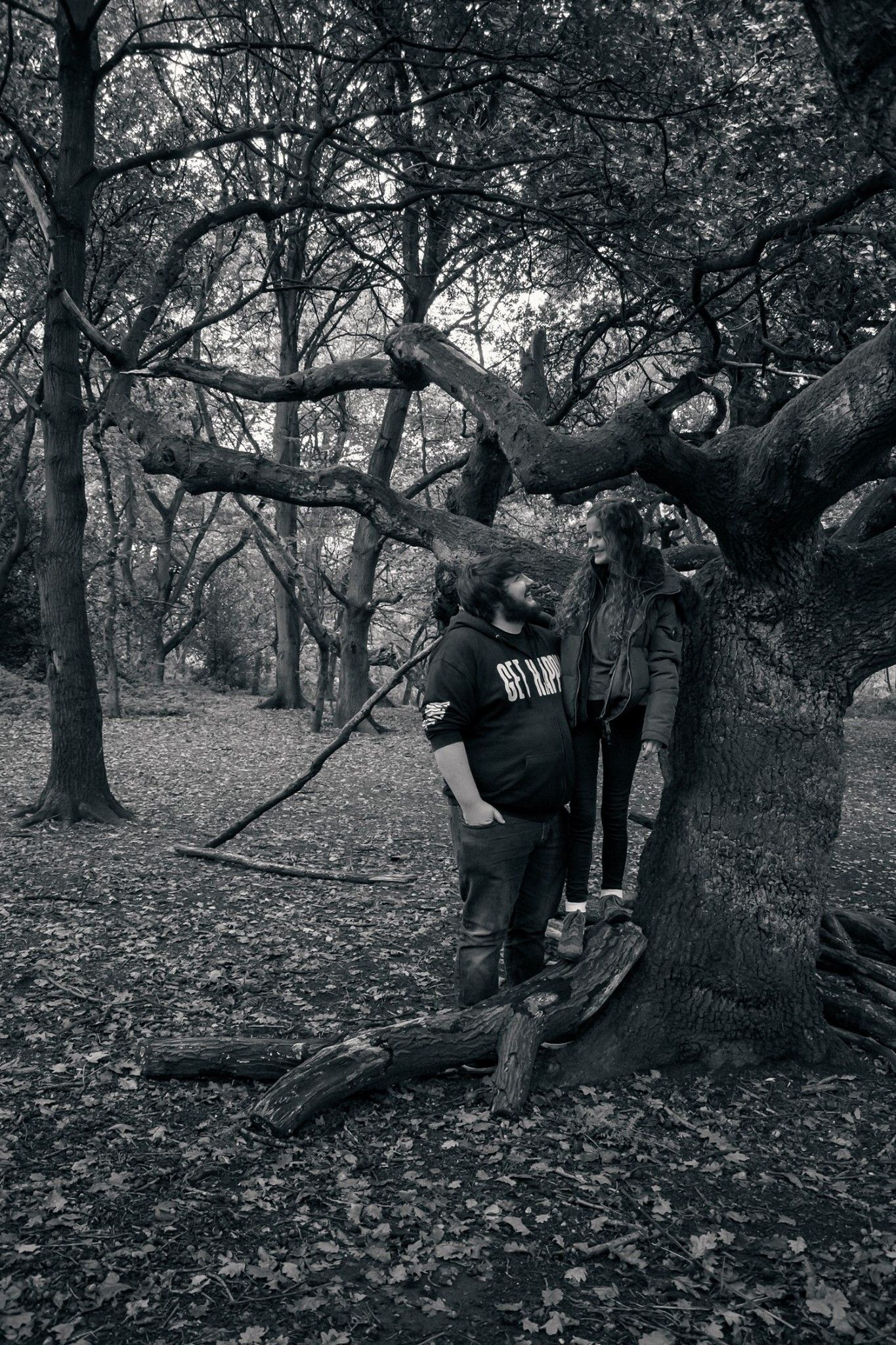 A couple posing together near a large tree in a moody black and white forest setting.