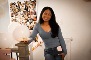 A person in a gray long-sleeve top and jeans poses casually against a wall decorated with photos.