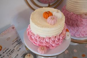 White cake with pink rosette details and small decorative pumpkins on white cake stand.