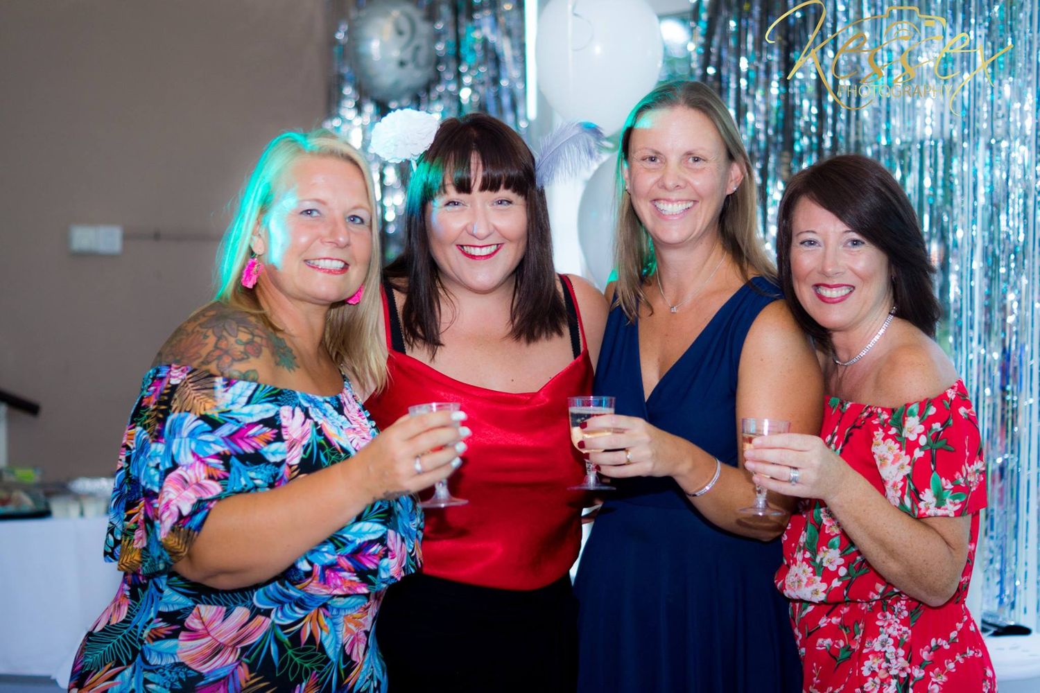 Four women in colorful dresses celebrate at a party with blue streamers in the background.