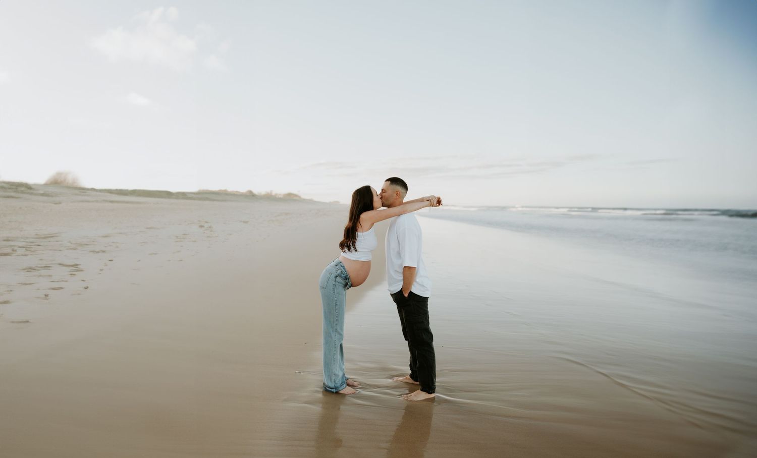 Couple embracing on beach at sunset wearing blue dress and dark clothing.
