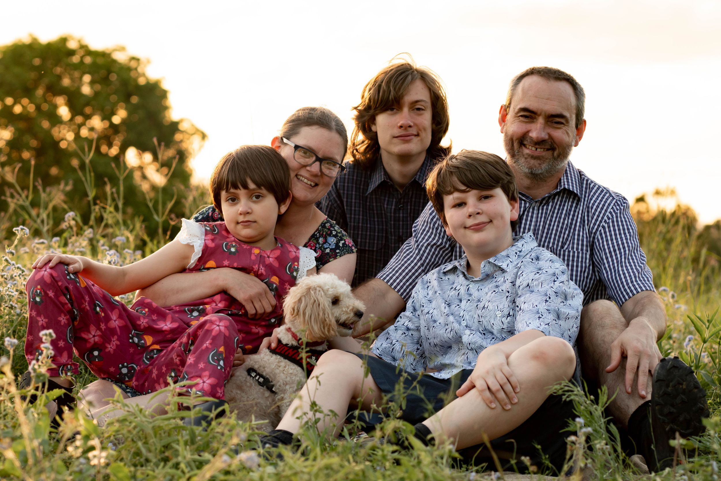 Family of five sit close together in a grassy field with their dog at sunset