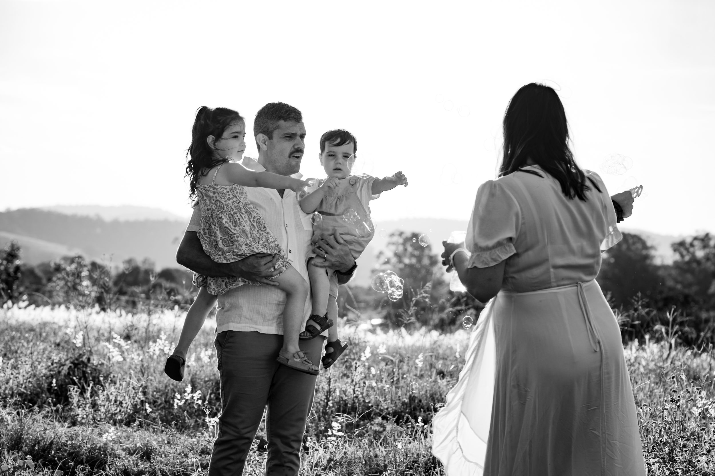 Black and white image of a young family; father carrying two children, while mother leans towards them blowing bubbles