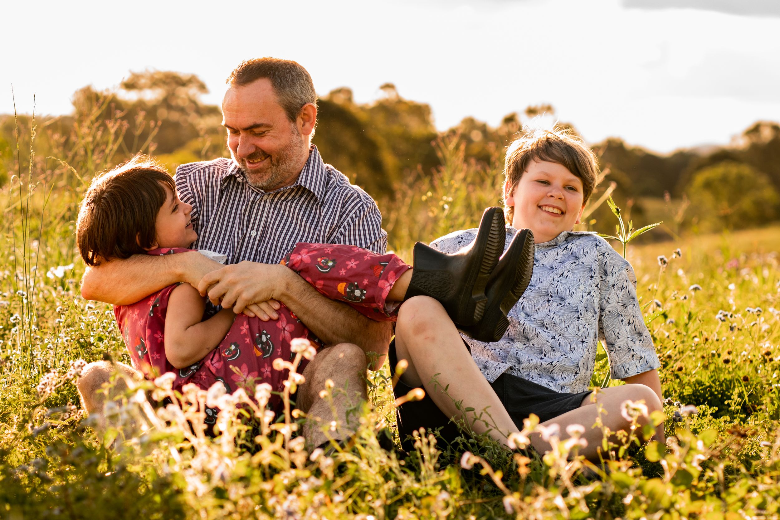 Father sits in long grass, cradling his daughter, while son sits laughing beside them.