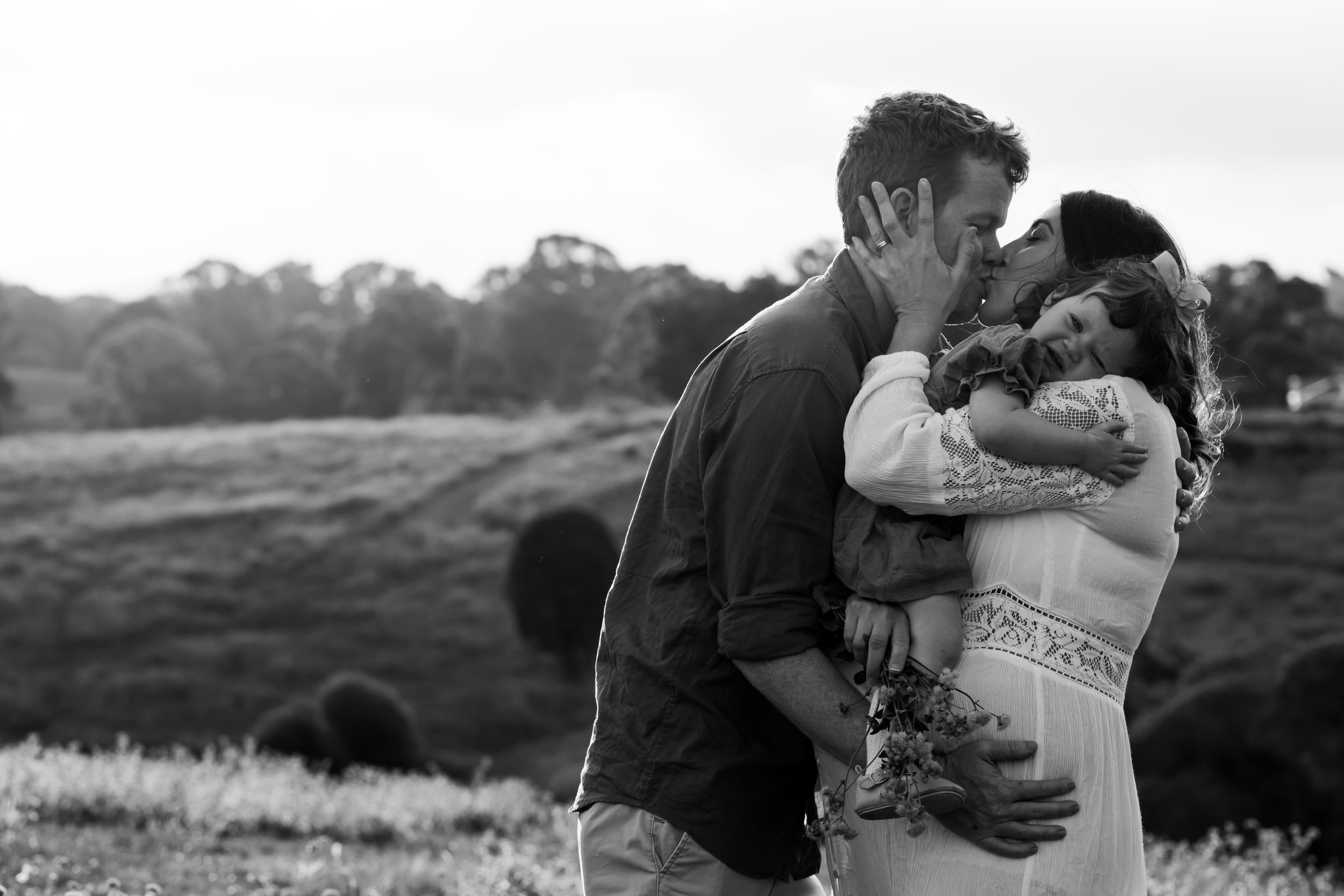 Black and white photograph of husband and wife stealing a kiss, while toddler cries on her mother's shoulder