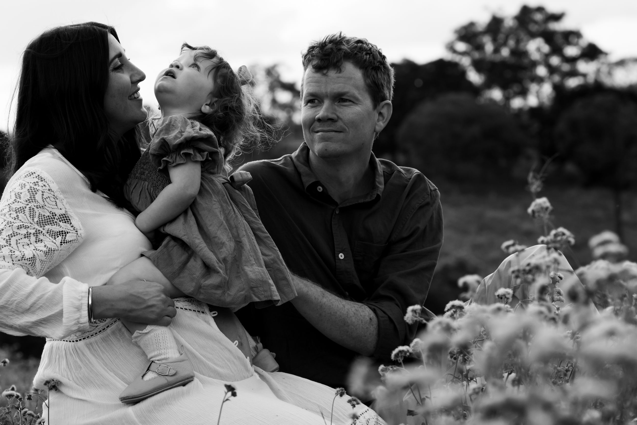 Father and mother seated amongst wildflowers. Toddler sits above mother's pregnant belly