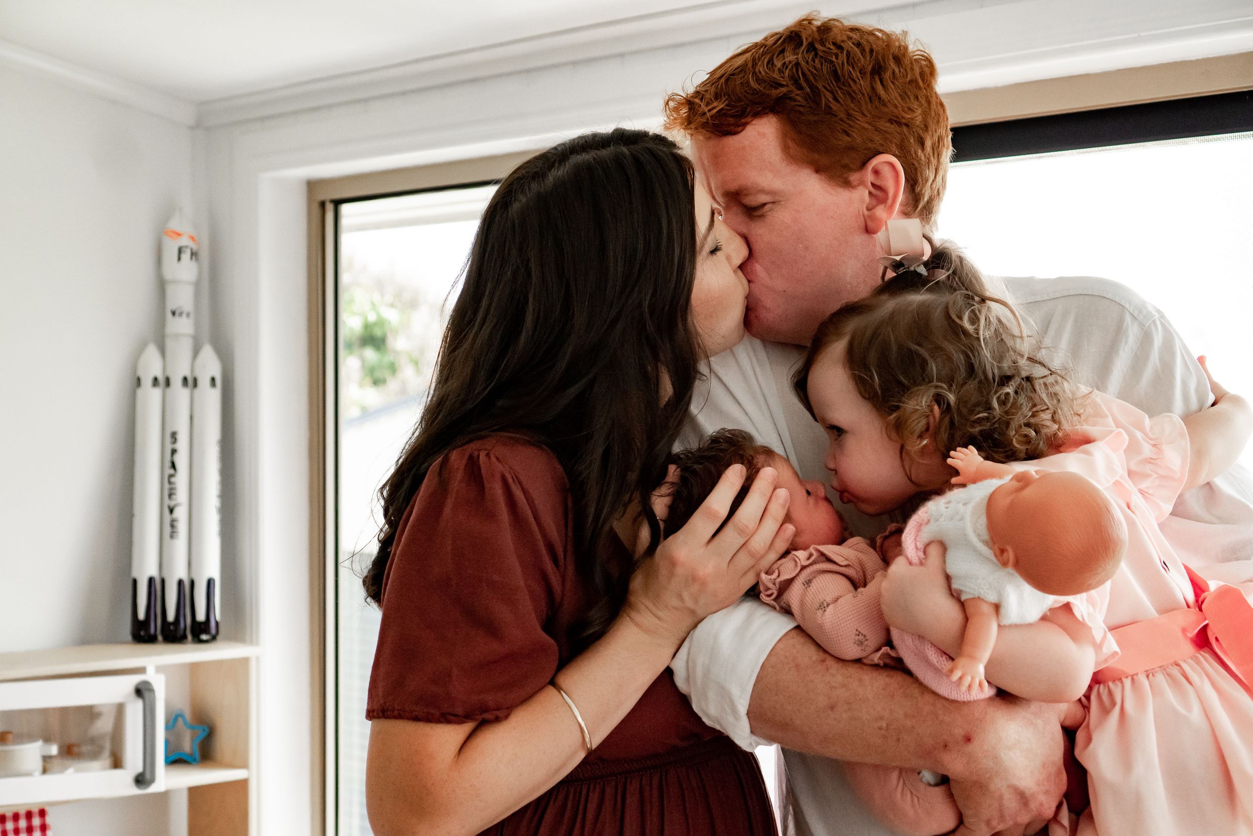 Husband and wife steal a kiss while cradling newborn baby and toddler. Toddler likewise leans in to kiss baby