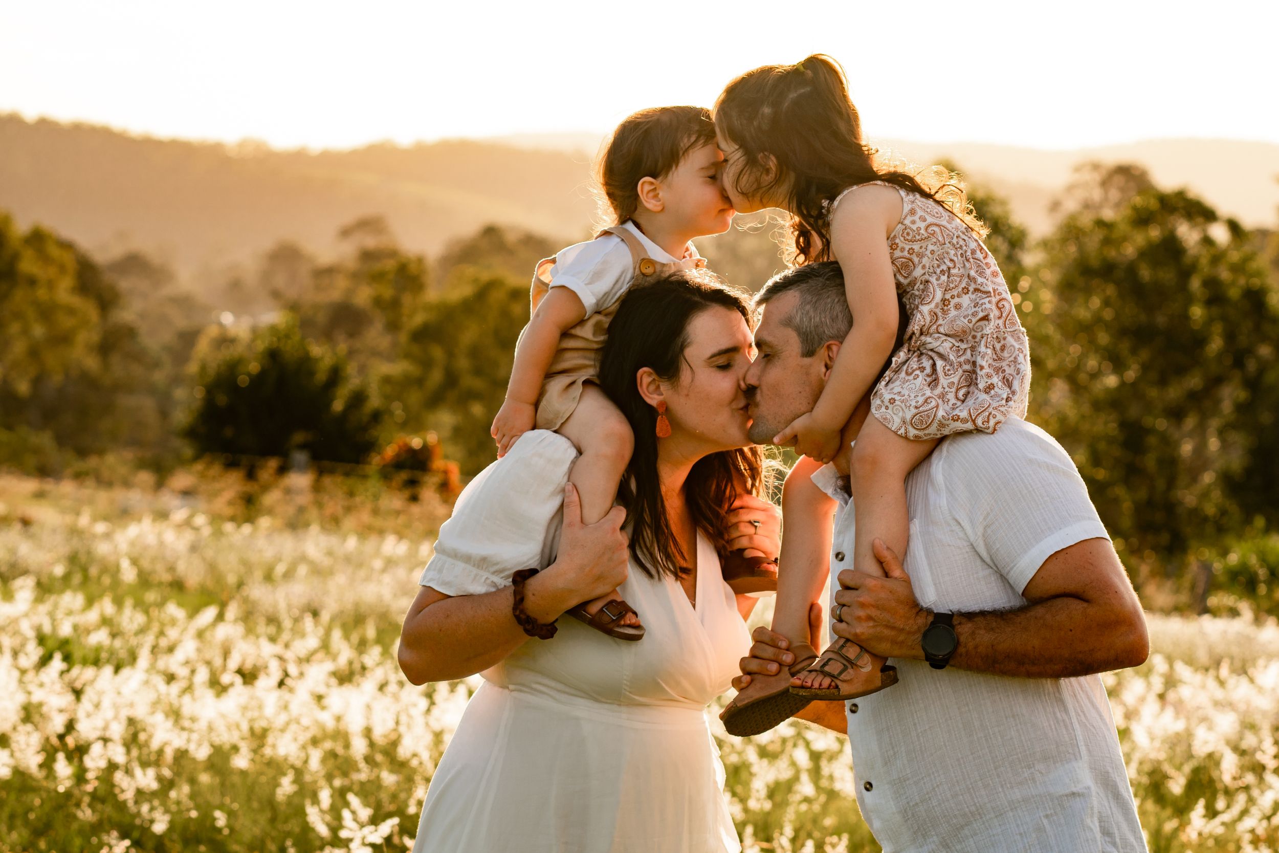 Mother and father kissing in a field at sunset, while carrying two young children on their shoulders