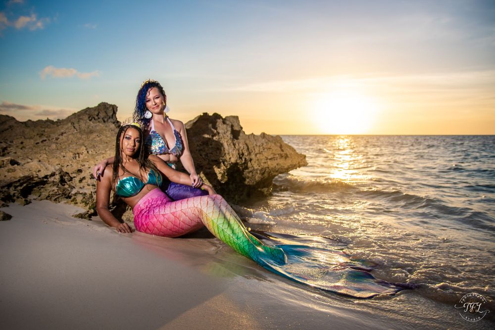 Mermaid performers in colorful tails pose on beach rocks at sunset with golden light reflecting on the ocean waves.