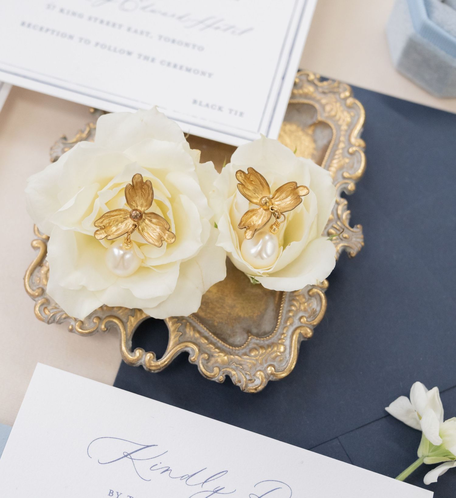 White roses with gold jewelry displayed on vintage ornate tray beside wedding invitation.