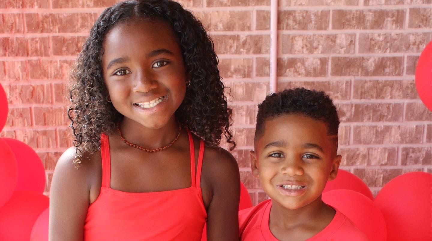 Two children wearing red outfits smile brightly against a brick wall background.