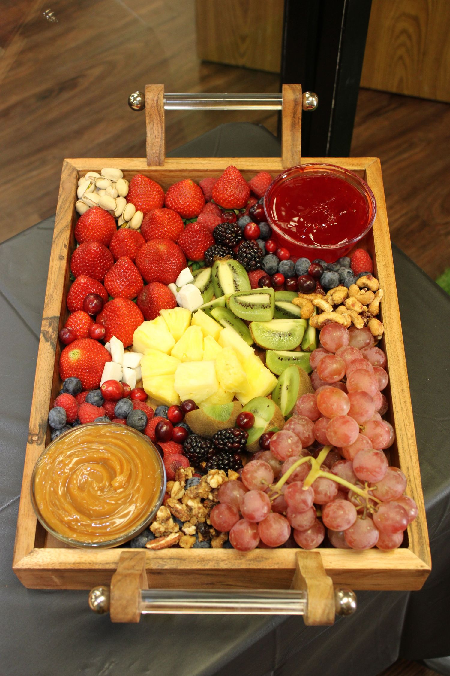 Wooden serving tray filled with fresh fruit, nuts, and dipping sauces including strawberries and grapes.